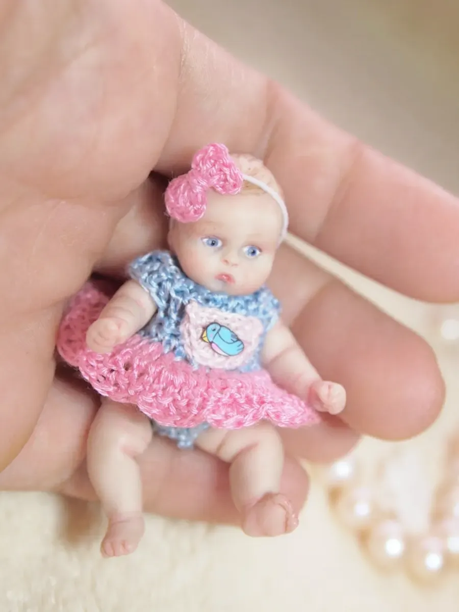 Adorable OOAK miniature baby doll with blue eyes and pink bow headband, wearing pink and blue crocheted dress with bird emblem, sitting in palm - by Mini Baby Dolls