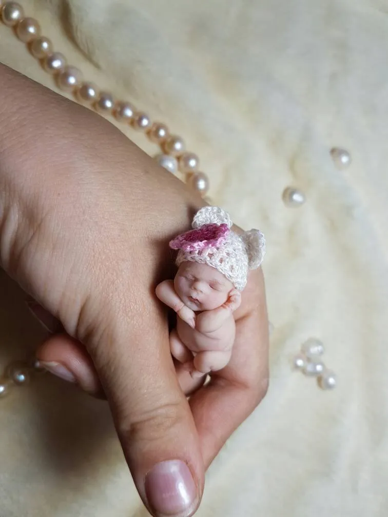 Adorable OOAK miniature baby doll wearing a pink and white crocheted bear-ear hat, resting peacefully in palm with a tiny pink teddy bear companion - by Mini Baby Dolls