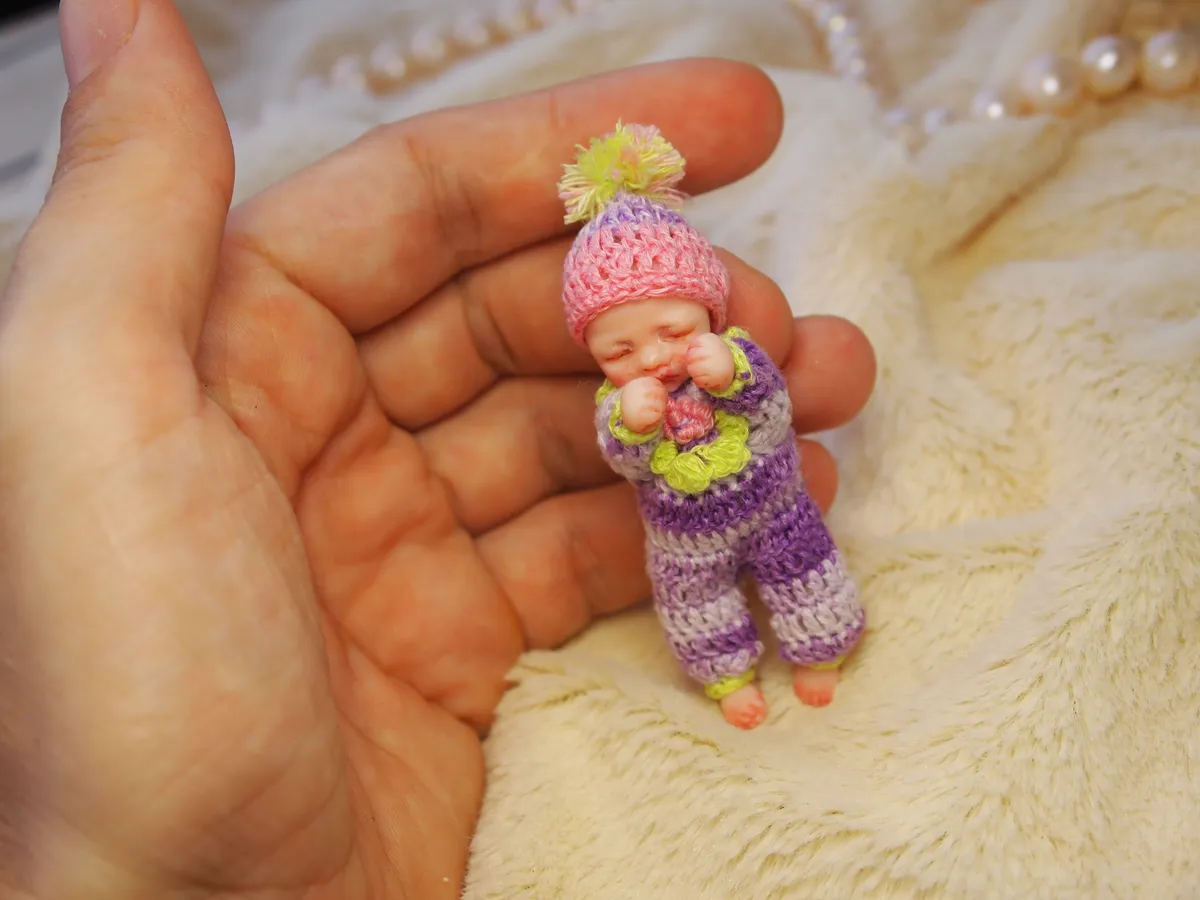 Cheerful OOAK miniature baby doll in colorful rainbow striped crocheted outfit with matching pom pom hat, holding a tiny flower, displayed in palm - by Mini Baby Dolls