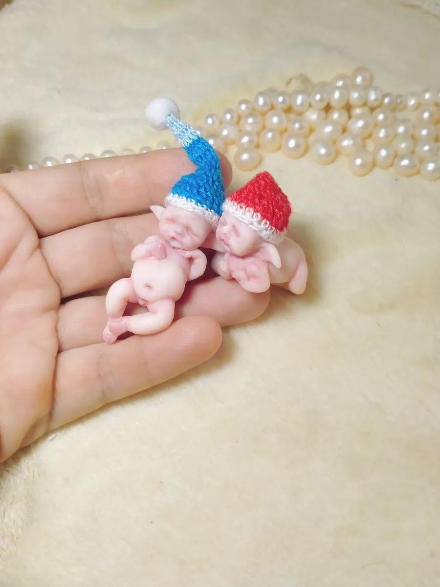 Two cuddling OOAK miniature baby dolls wearing matching red and blue crocheted hats, displayed together with pearl beads - by Mini Baby Dolls