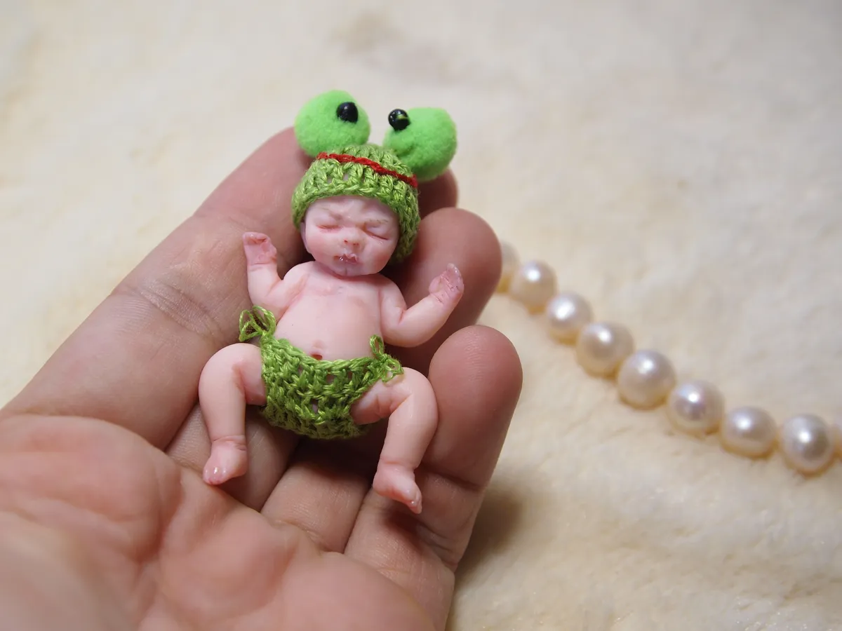 Sleeping OOAK miniature baby doll in green crocheted frog hat with googly eyes and matching diaper, relaxing in palm - by Mini Baby Dolls