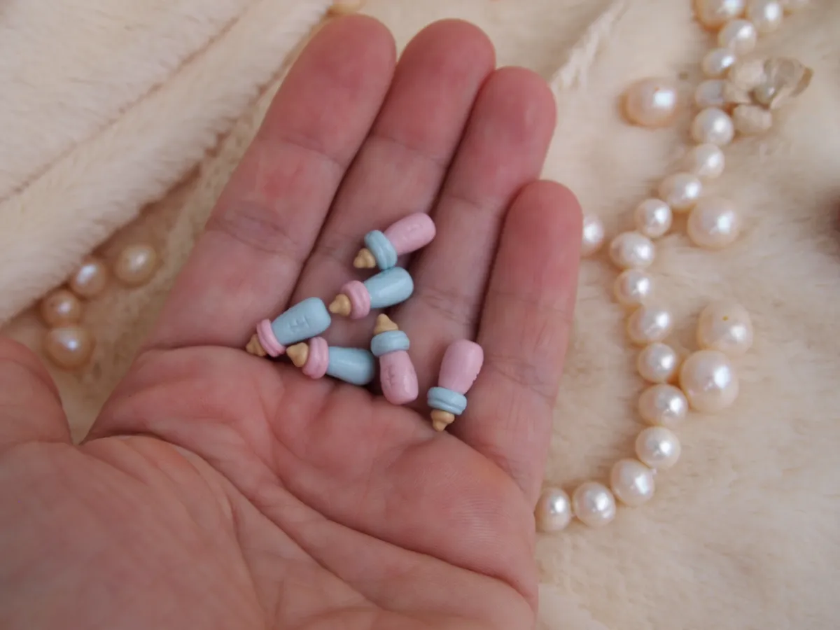 Handcrafted miniature baby bottle accessories in pastel blue and tan colors, displayed in palm with pearl beads - by Mini Baby Dolls