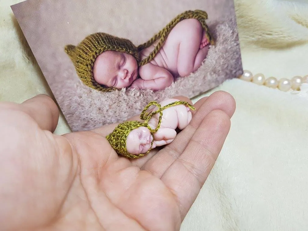 Sleeping OOAK miniature baby doll in olive green crocheted outfit with matching bonnet, displayed with printed photo reference in background - by Mini Baby Dolls