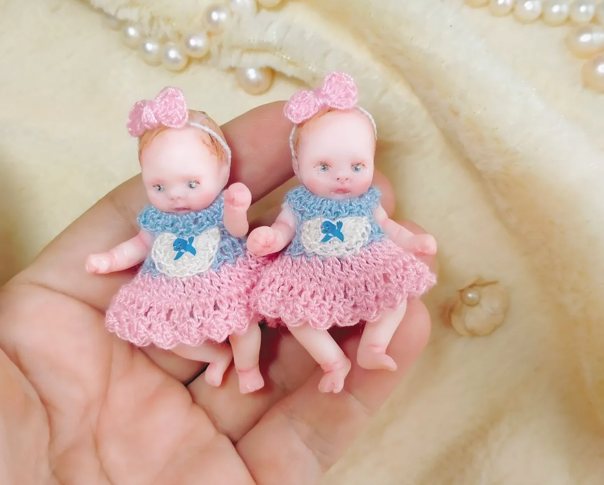 Adorable matching pair of OOAK miniature twin dolls in handmade pink and blue crocheted dresses with bow headbands, displayed together in palm - by Mini Baby Dolls