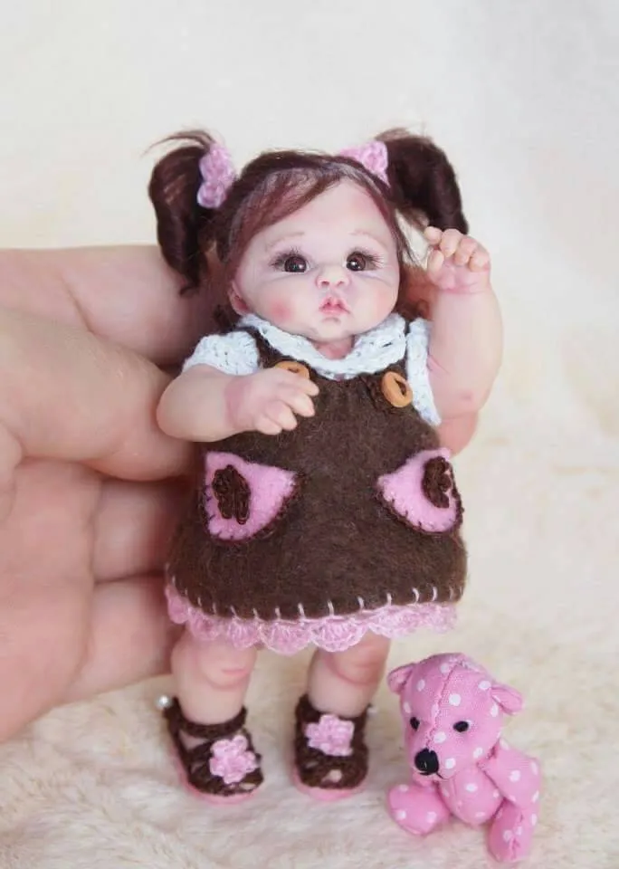Detailed OOAK miniature baby doll with dark pigtails and pink bows, wearing a brown felt dress with pink heart details and crocheted collar, posing with a tiny pink teddy bear - by Mini Baby Dolls