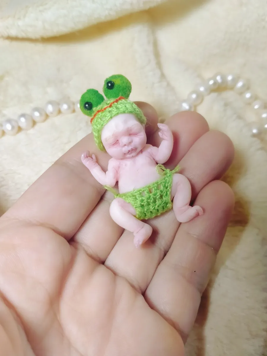 Playful OOAK miniature baby doll in green crocheted frog costume with googly eyes, displayed in palm with pearl beads - by Mini Baby Dolls