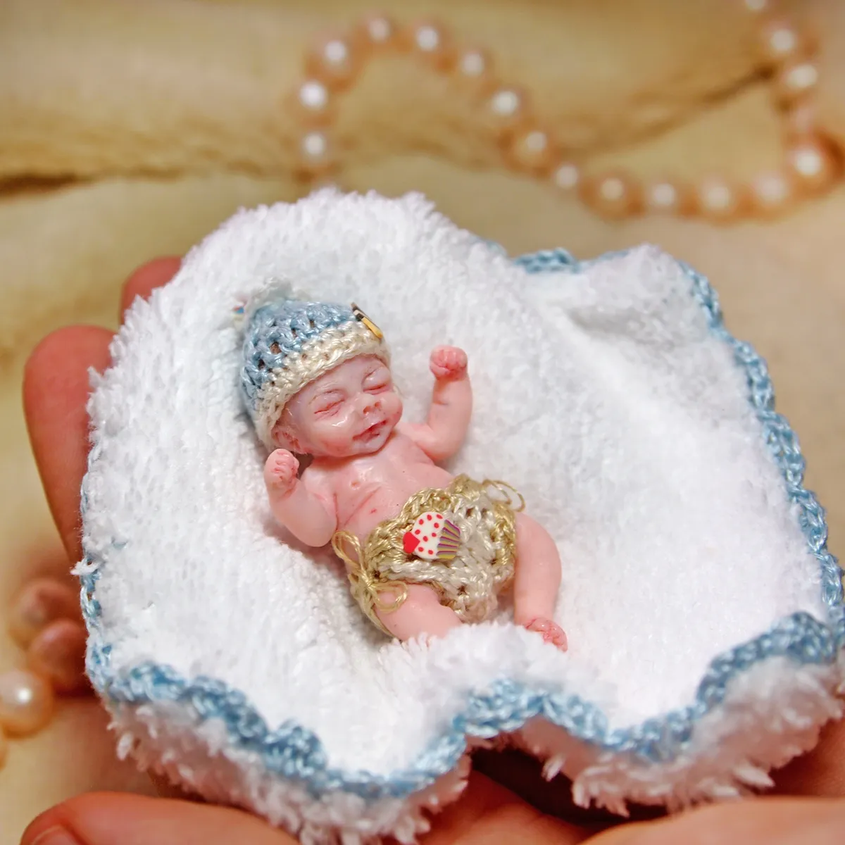 Regal OOAK miniature baby doll wearing a golden crown and delicate gold lace outfit, resting on handmade white crocheted blanket with blue trim, displayed with pearl beads - by Mini Baby Dolls