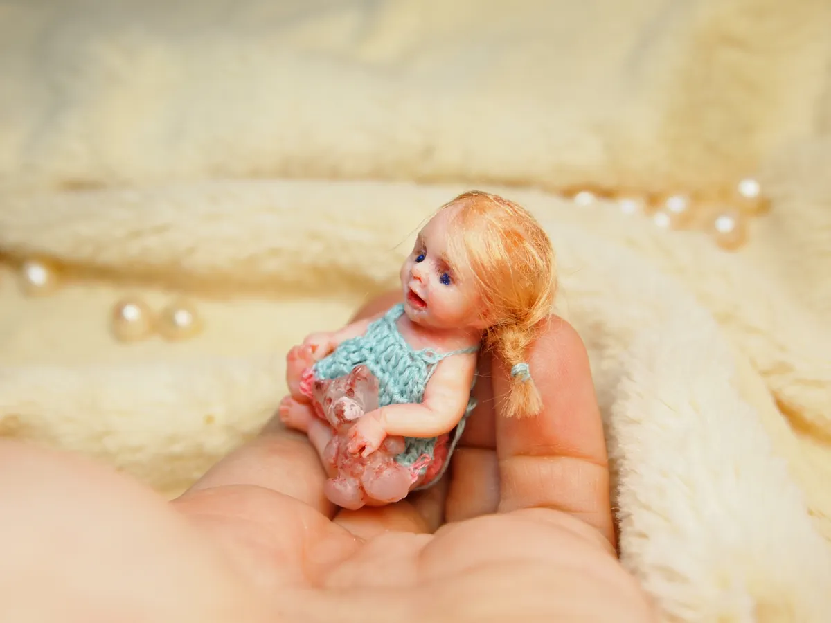 Detailed OOAK miniature baby doll with orange-red hair in pigtails, wearing a turquoise crocheted outfit, sitting in palm with pearl beads - by Mini Baby Dolls