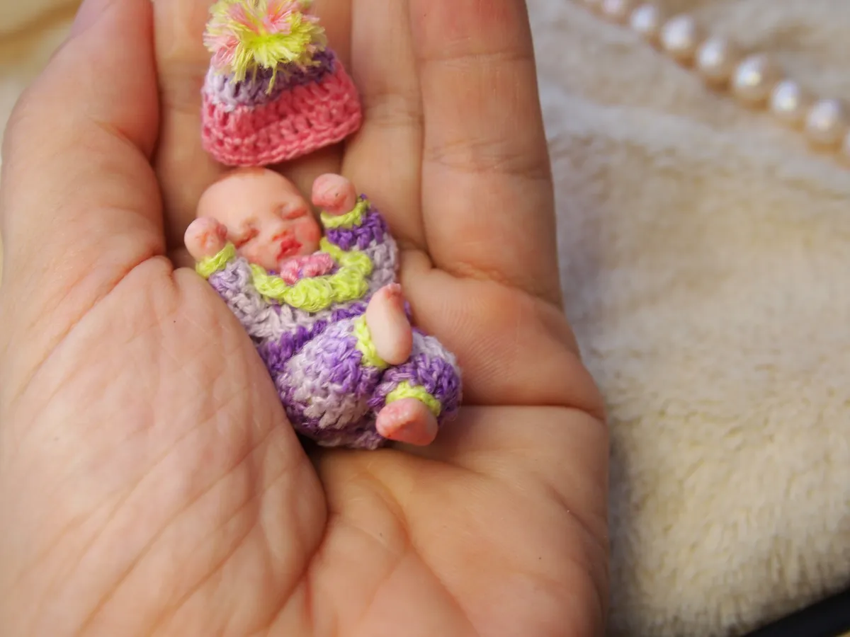 Sleeping OOAK miniature baby doll in colorful handmade crocheted outfit with purple, yellow and green stripes and matching rainbow hat, held in palm - by Mini Baby Dolls