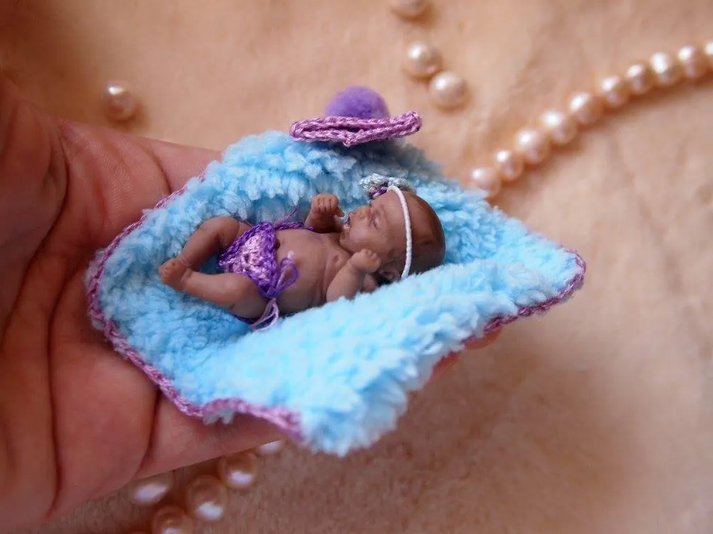 Sleeping OOAK miniature baby doll wearing a purple crocheted hat, resting on a handmade blue crocheted blanket or pillow, displayed with pearl beads - by Mini Baby Dolls