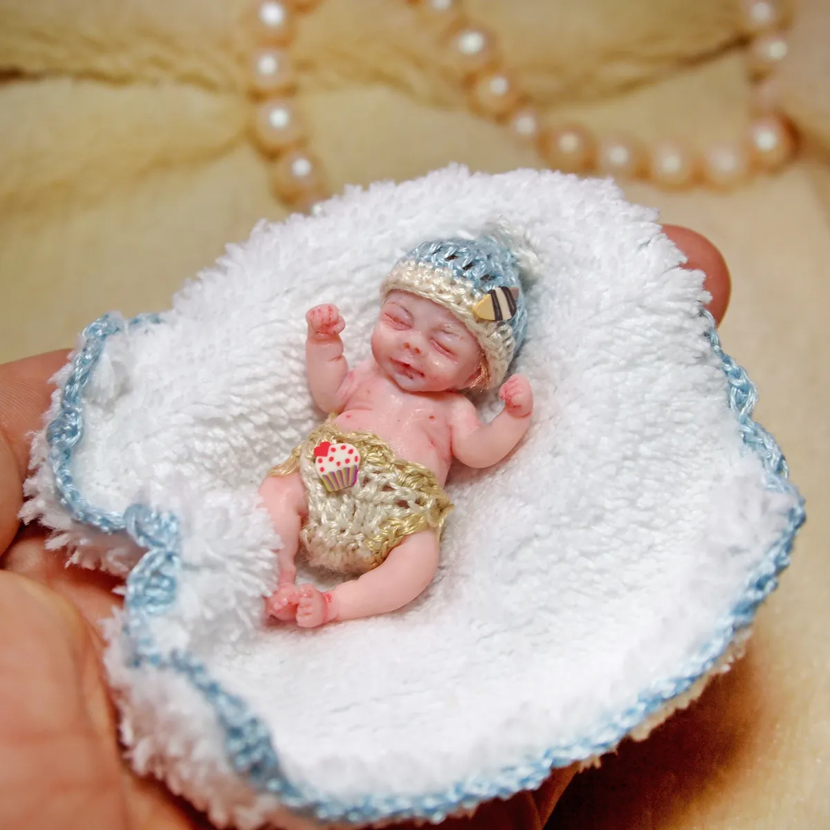 Sleeping OOAK miniature baby doll resting on a handmade white crocheted blanket with delicate blue trim, displayed with pearl beads - by Mini Baby Dolls