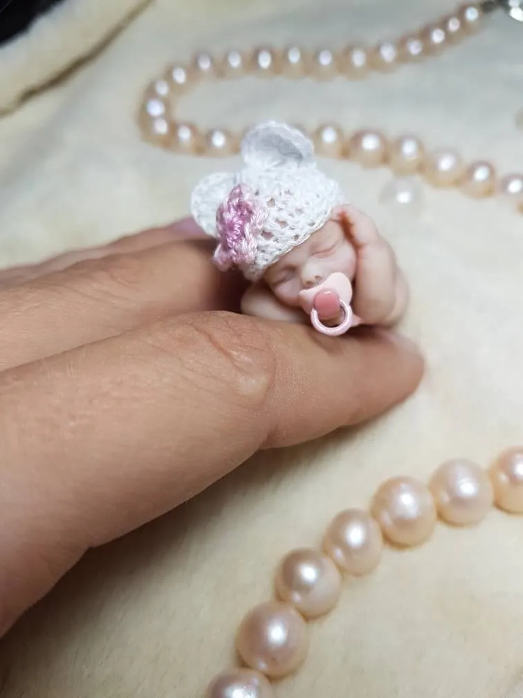 Sleeping OOAK miniature baby doll in white crocheted outfit with purple ribbon bow, holding a tiny pacifier, displayed with pearl beads - by Mini Baby Dolls