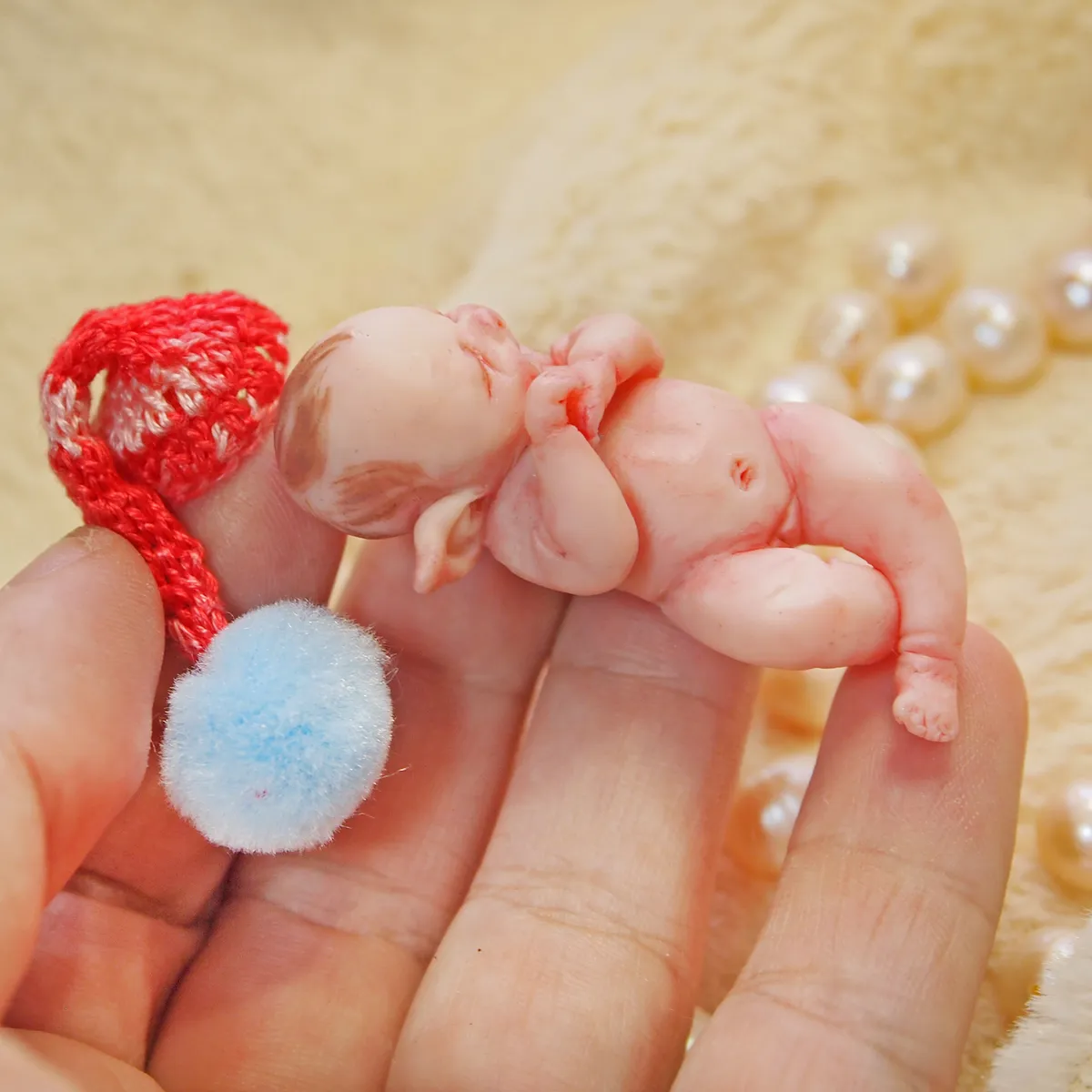 Sleeping OOAK miniature elf baby doll with pointed ears wearing a bright red crocheted hat, cuddling a blue pom pom, displayed with pearl beads - by Mini Baby Dolls