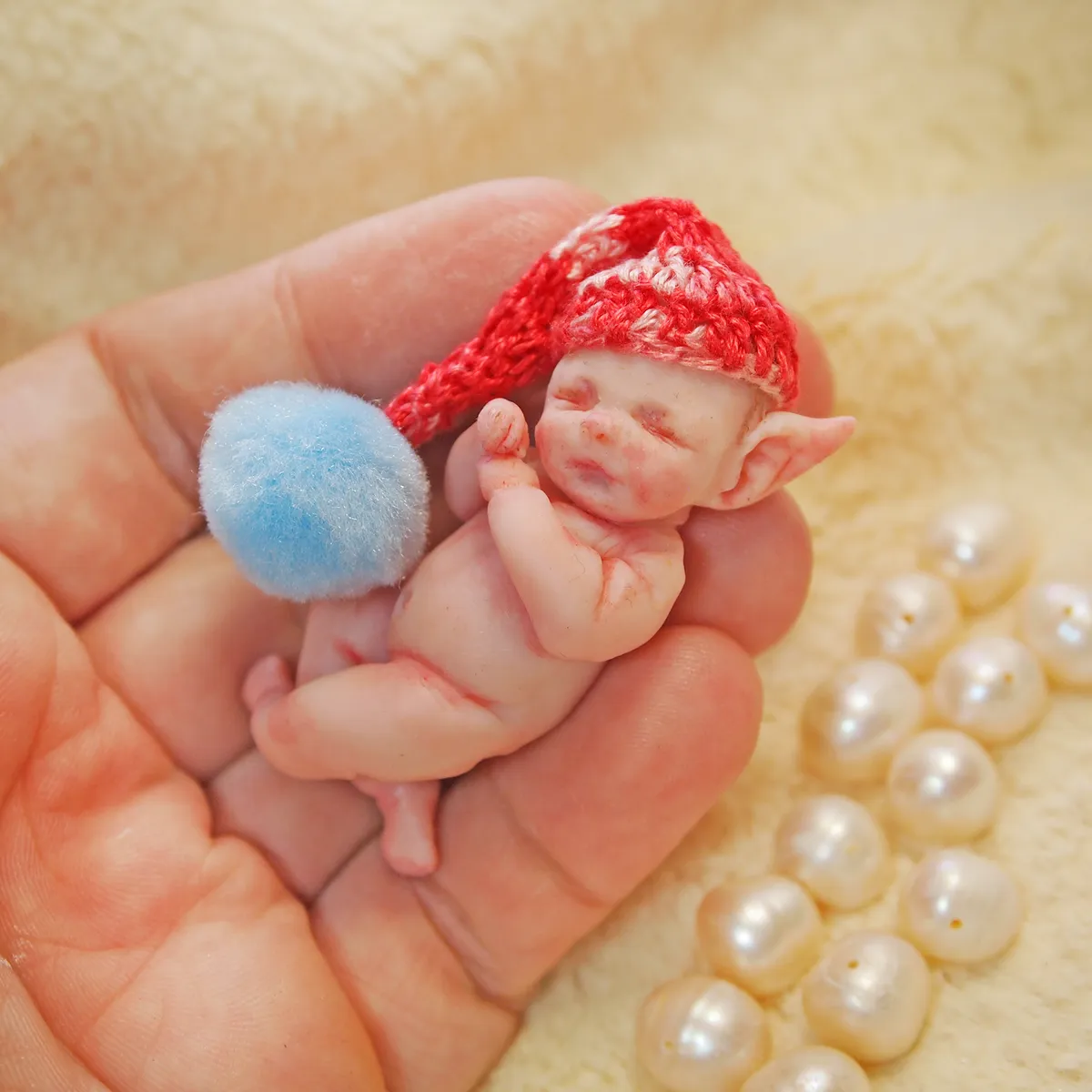 Sleeping OOAK miniature elf baby doll with pointed ears wearing a bright red crocheted gnome hat, cuddling a blue pom pom, held gently in palm - by Mini Baby Dolls
