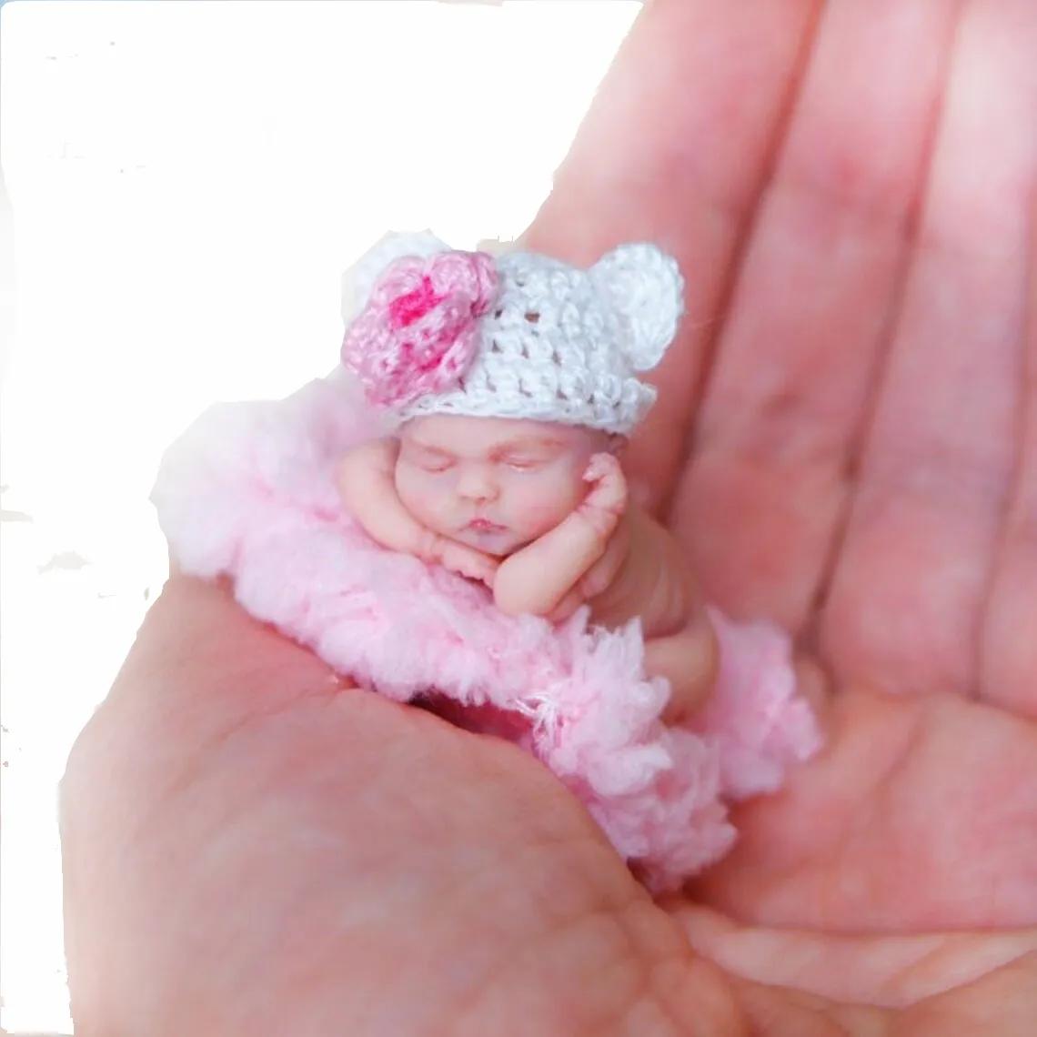 Handcrafted sleeping miniature baby doll in fluffy pink outfit with adorable pink bear hat, held gently in palm showing its tiny realistic scale - by Mini Baby Dolls