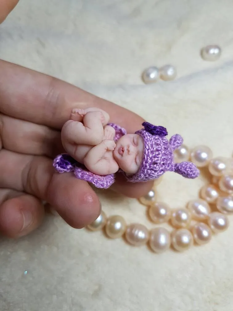 Tiny sleeping OOAK miniature baby doll in purple crocheted outfit, displayed inside a circle of elegant pearl beads showcasing miniature scale - by Mini Baby Dolls