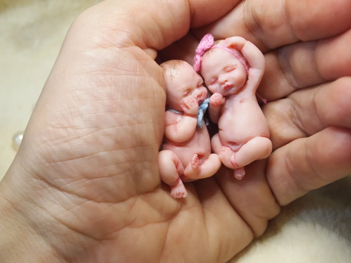Adorable twin OOAK miniature baby dolls cuddling together in palm, one wearing pink bow headband - by Mini Baby Dolls