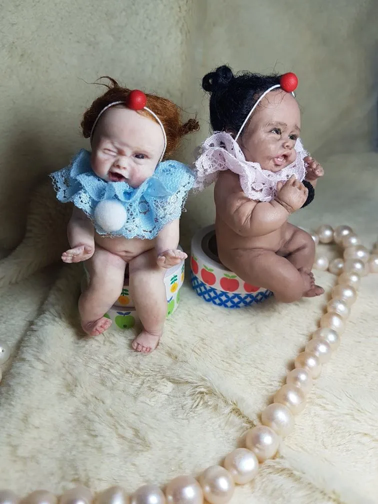 Two expressive OOAK miniature baby dolls with playful crying faces, wearing ladybug-style antennae hats - one in blue lace dress, one in pink polka dots, sitting on colorful cushions - by Mini Baby Dolls