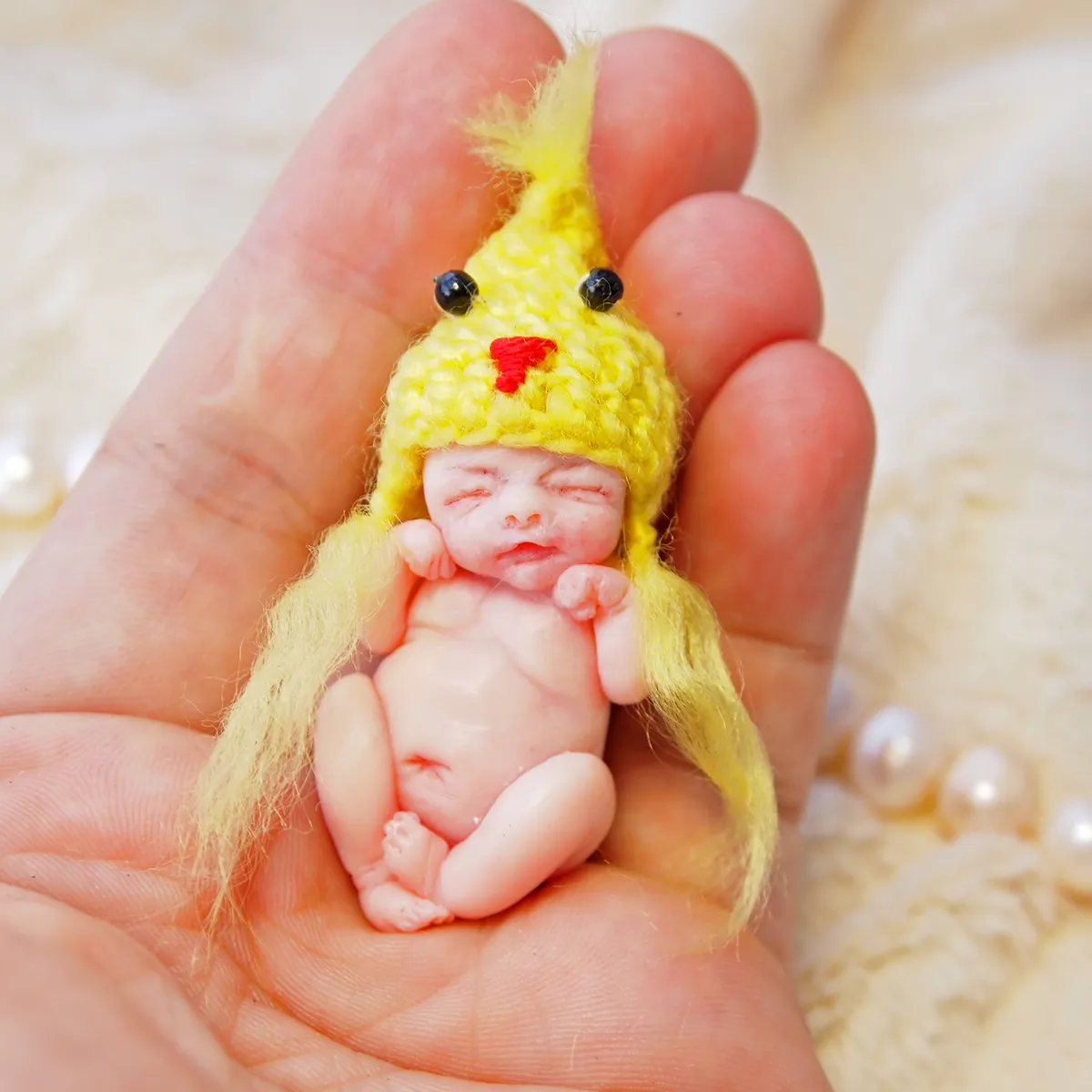 Happy OOAK miniature baby doll in yellow crocheted chick hat and feathery yellow outfit, displayed in palm with pearl beads - by Mini Baby Dolls